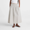 A woman modeling a white short-sleeve t-shirt and a long, pleated, smocked-waist maxi skirt in a light beige and white stripe pattern, paired with black sandals, against a plain white studio background.