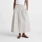 A woman modeling a white short-sleeve t-shirt and a long, pleated, smocked-waist maxi skirt in a light beige and white stripe pattern, paired with black sandals, against a plain white studio background.