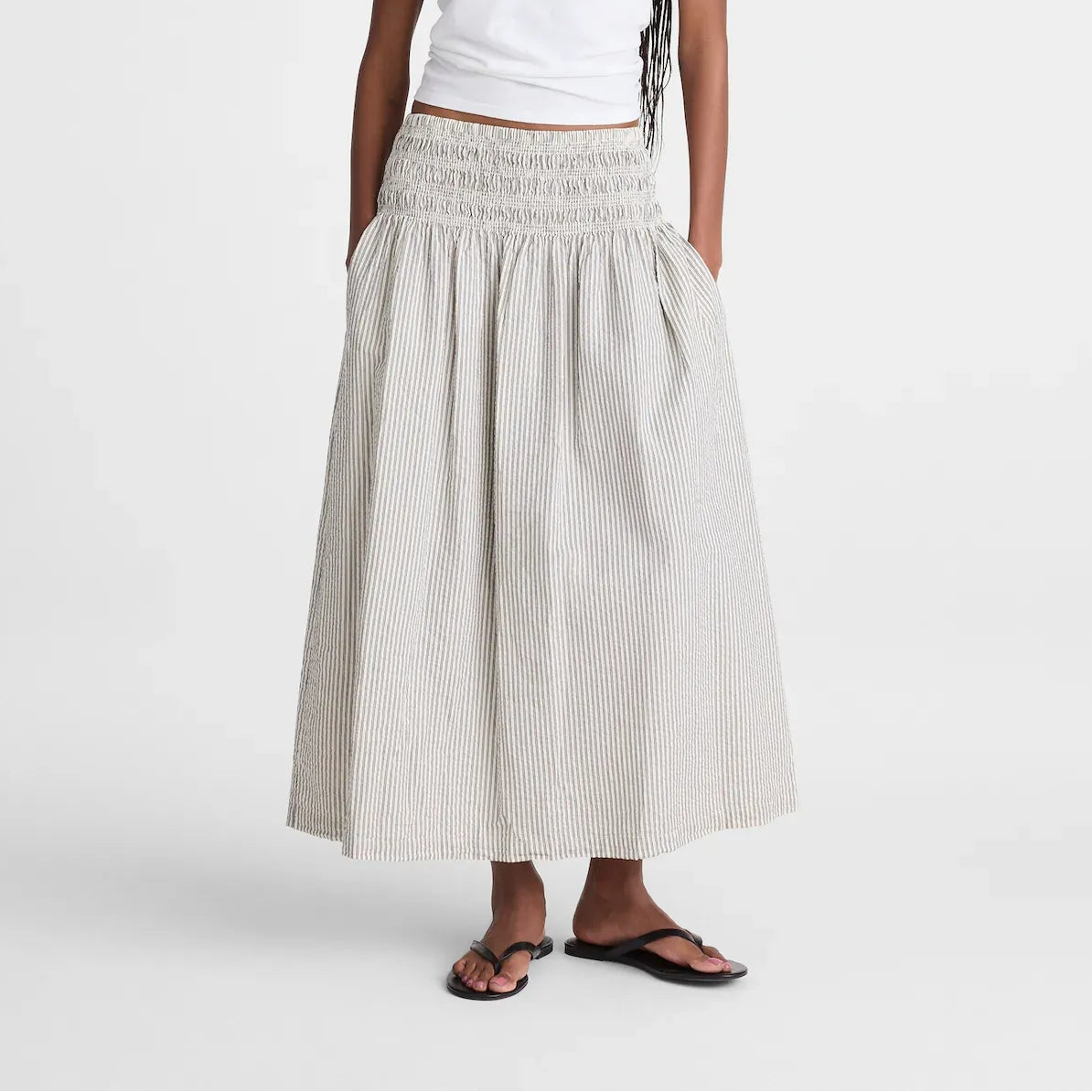 A woman modeling a white short-sleeve t-shirt and a long, pleated, smocked-waist maxi skirt in a light beige and white stripe pattern, paired with black sandals, against a plain white studio background.