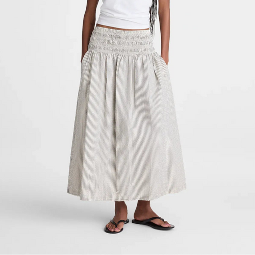A woman modeling a white short-sleeve t-shirt and a long, pleated, smocked-waist maxi skirt in a light beige and white stripe pattern, paired with black sandals, against a plain white studio background.