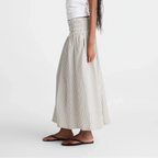 A woman modeling a white short-sleeve t-shirt and a high-waisted, pleated light beige maxi skirt with a smocked waistband, paired with black flip-flops against a plain white studio background.