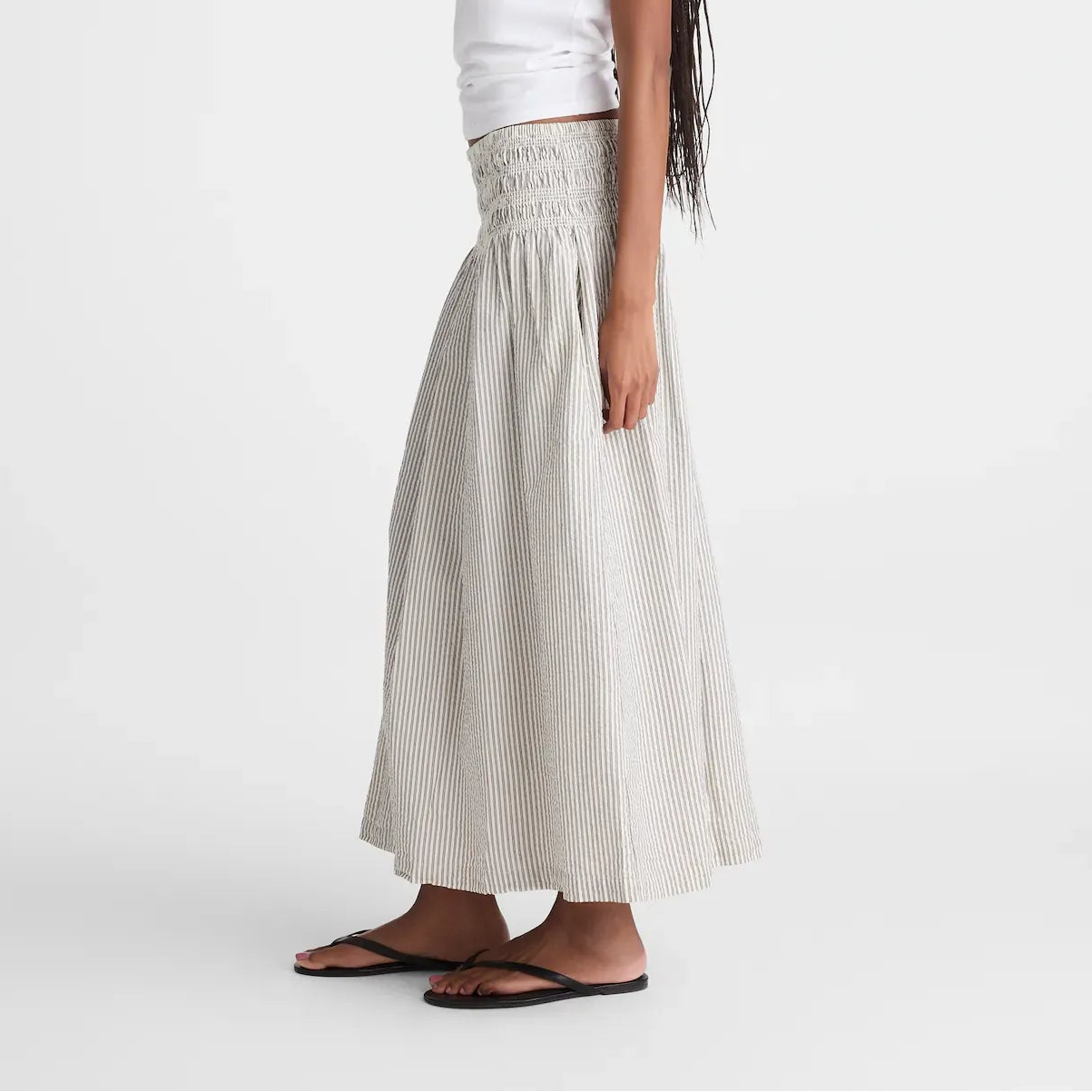 A woman modeling a white short-sleeve t-shirt and a high-waisted, pleated light beige maxi skirt with a smocked waistband, paired with black flip-flops against a plain white studio background.