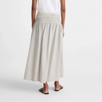 A woman shown from the back wearing a white short-sleeve t-shirt and a light beige smocked maxi skirt with subtle vertical stripes, paired with black flat sandals, against a plain white studio background.