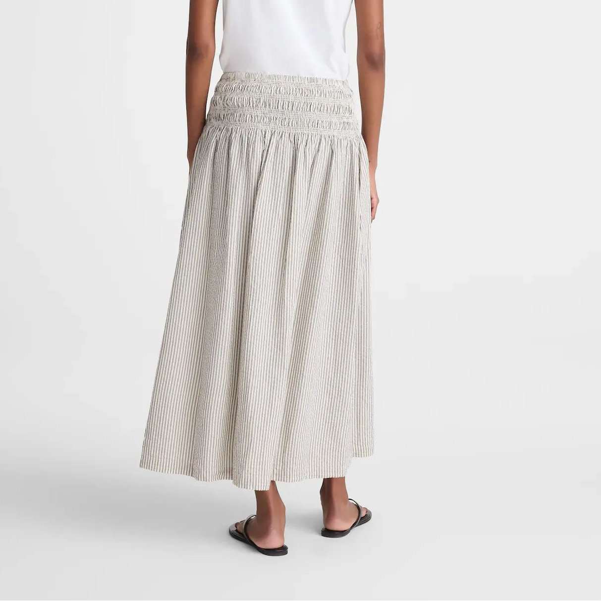 A woman shown from the back wearing a white short-sleeve t-shirt and a light beige smocked maxi skirt with subtle vertical stripes, paired with black flat sandals, against a plain white studio background.