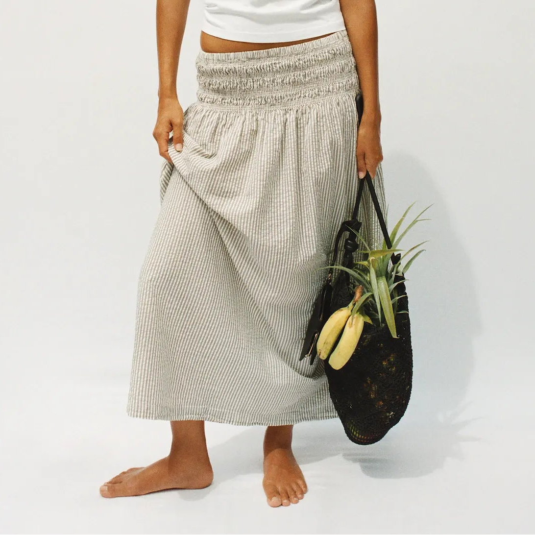 A woman stands barefoot against a plain light gray studio background, wearing a white short-sleeve cropped T-shirt and a loose light beige patterned maxi skirt. She holds a black woven tote bag with a plant in it.