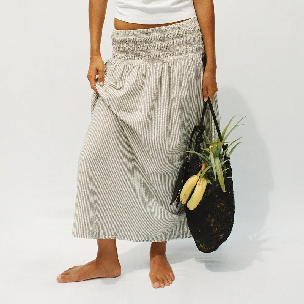 A woman stands barefoot against a plain light gray studio background, wearing a white short-sleeve cropped T-shirt and a loose light beige patterned maxi skirt. She holds a black woven tote bag with a plant in it.