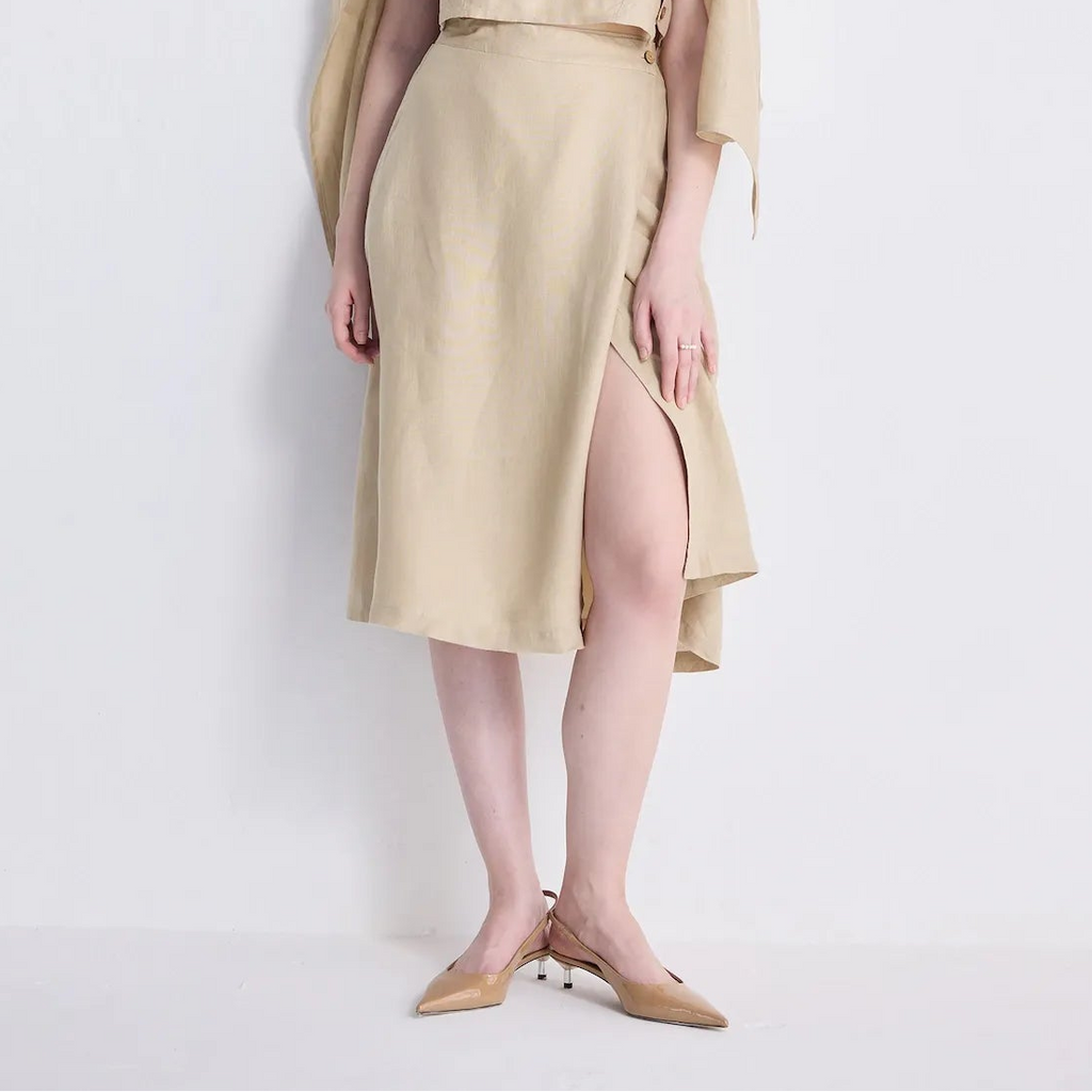 A woman models a beige linen two-piece set consisting of a sleeveless crop top and matching midi skirt with a high side slit, styled with a matching beige beret and draped cardigan, against a plain white studio background.