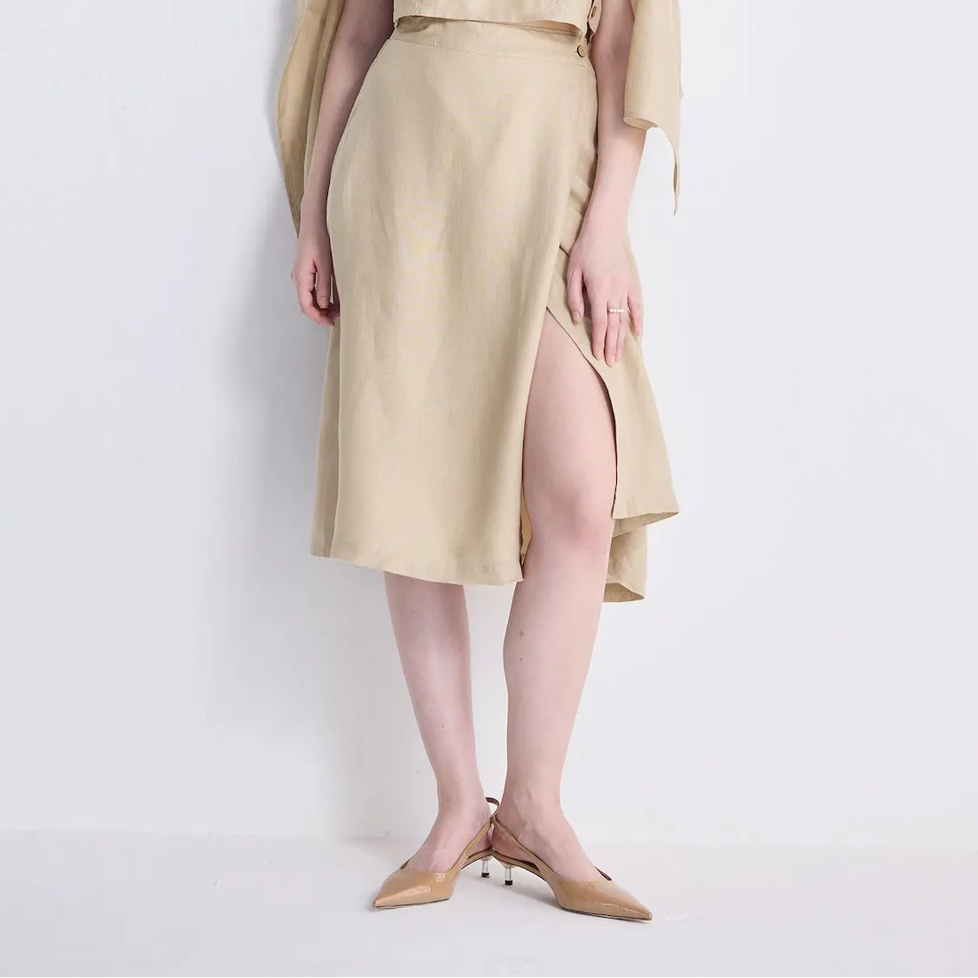 A woman models a beige linen two-piece set consisting of a sleeveless crop top and matching midi skirt with a high side slit, styled with a matching beige beret and draped cardigan, against a plain white studio background.