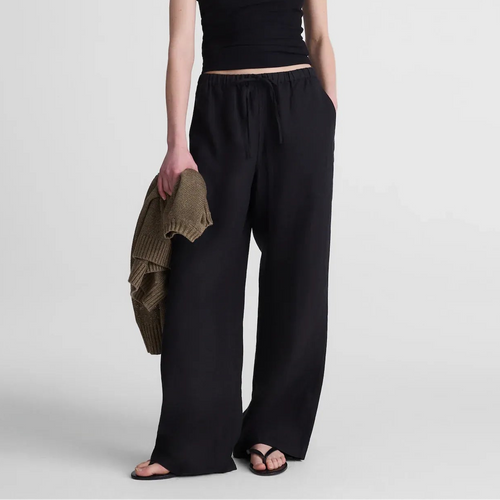 A model stands against a light gray studio background wearing a black spaghetti-strap crop top and matching black wide-leg pants, holding a folded olive-brown knit garment in one hand.