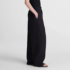 A woman modeling a black sleeveless maxi dress with wide-leg pants and side pockets, standing in profile against a light gray studio background.