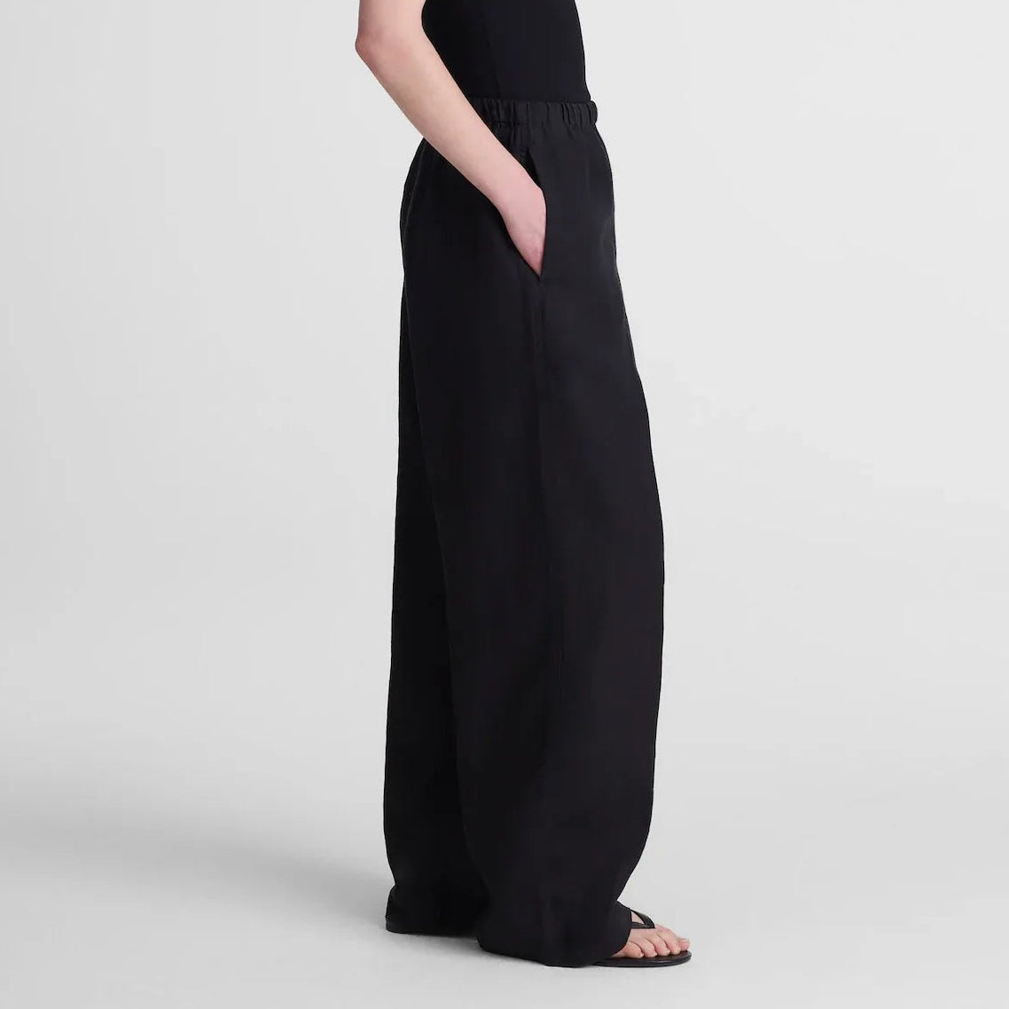 A woman modeling a black sleeveless maxi dress with wide-leg pants and side pockets, standing in profile against a light gray studio background.
