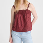 A woman modeling a burgundy red sleeveless camisole top with a scalloped neckline and subtle tonal floral embroidery, paired with light blue jeans, against a plain light gray background.