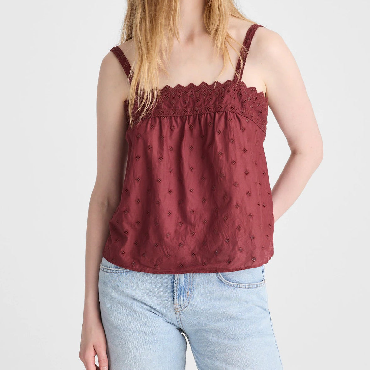 A woman modeling a burgundy red sleeveless camisole top with a scalloped neckline and subtle tonal floral embroidery, paired with light blue jeans, against a plain light gray background.