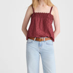 A woman modeling a burgundy sleeveless top with a scalloped neckline and subtle polka dot pattern, paired with light-wash wide-leg jeans and brown sandals, against a plain light gray studio background.