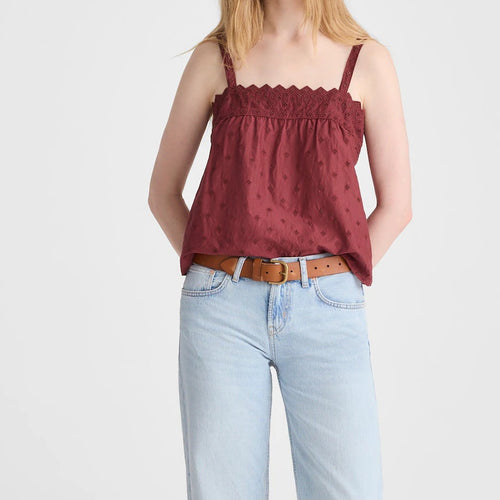 A woman modeling a burgundy sleeveless top with a scalloped neckline and subtle polka dot pattern, paired with light-wash wide-leg jeans and brown sandals, against a plain light gray studio background.