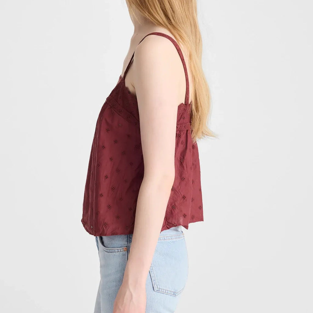 A woman with long blonde hair is shown in profile wearing a burgundy camisole top with a subtle pattern and light blue jeans, photographed against a solid light gray background.
