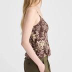 A woman in side profile wearing a brown floral print camisole top and olive green pants, photographed against a light gray studio background.