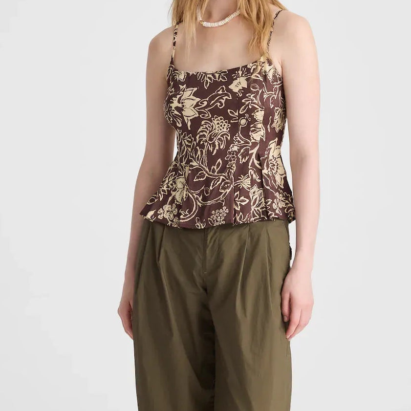 A woman stands against a light gray studio background wearing a sleeveless floral camisole top with a brown and beige print, loose olive green pants, and black sandals.