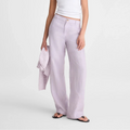 A model wearing a white ribbed tank top, light lavender wide-leg linen trousers, and black sunglasses, holding a matching lavender linen shirt against a plain light gray studio background.