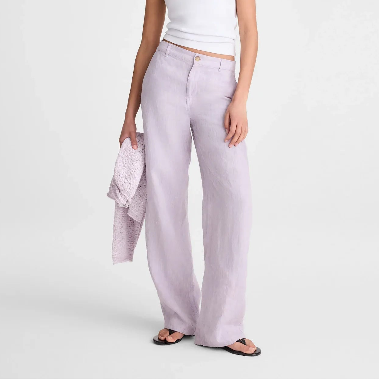 A model wearing a white ribbed tank top, light lavender wide-leg linen trousers, and black sunglasses, holding a matching lavender linen shirt against a plain light gray studio background.