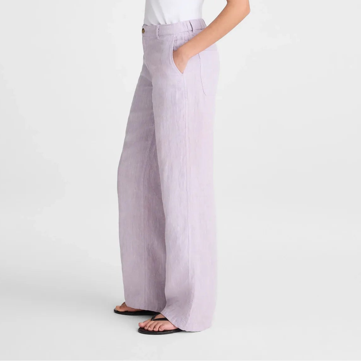 A woman in a white tank top and light lavender wide-leg trousers stands in profile against a clean white studio background.