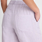 Close-up of a person wearing light lavender wide-leg trousers with an elastic waistband, side pockets, and a back pocket, paired with a white top against a plain light background.