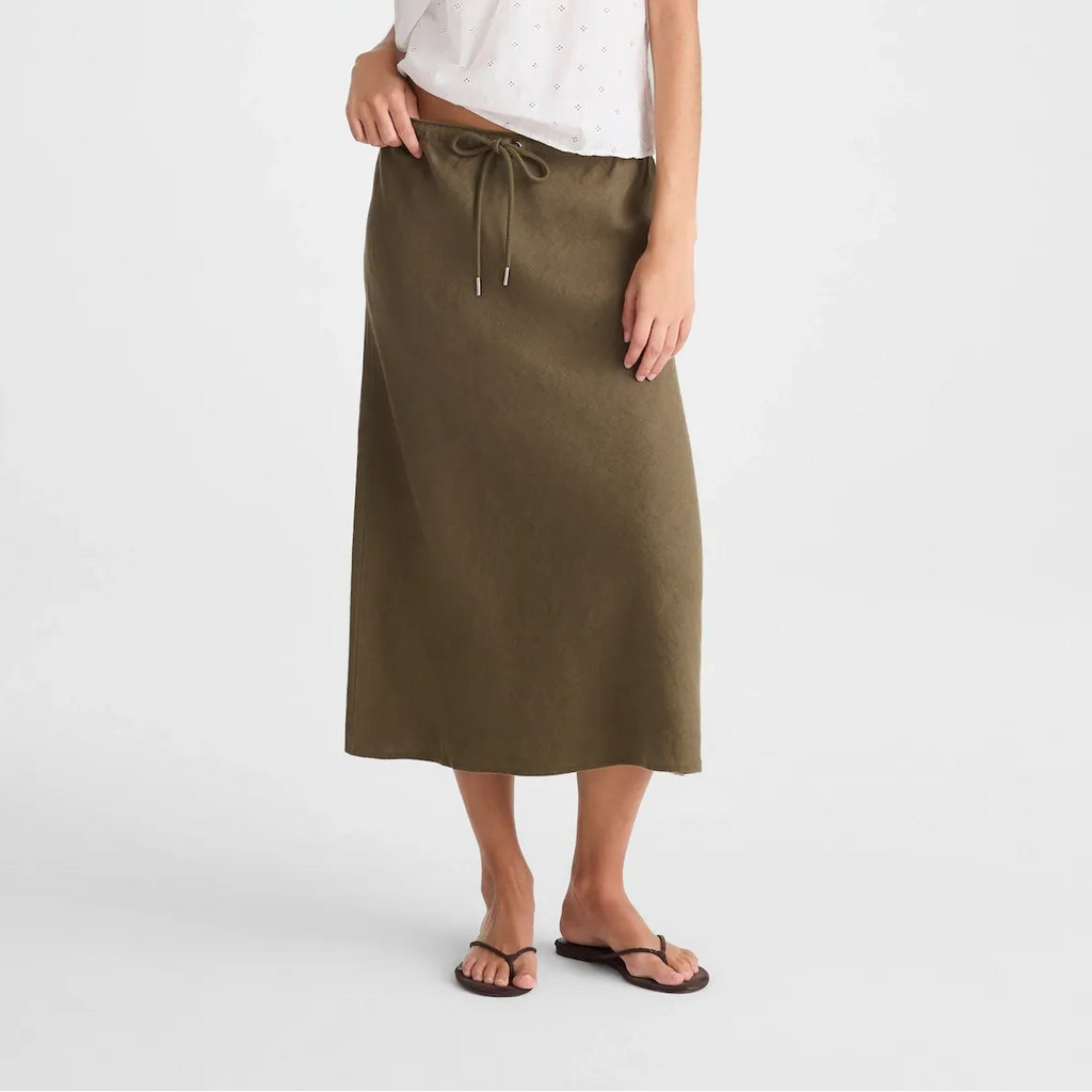 A woman modeling a white sleeveless eyelet camisole top with lace trim and a high-waisted olive green midi skirt, styled with a beaded necklace and black sandals against a light gray studio background.