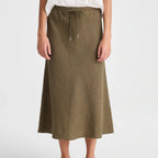 A woman wearing an olive green midi skirt with a drawstring waist, paired with a white top and black flip-flop sandals, shown against a light gray studio background.