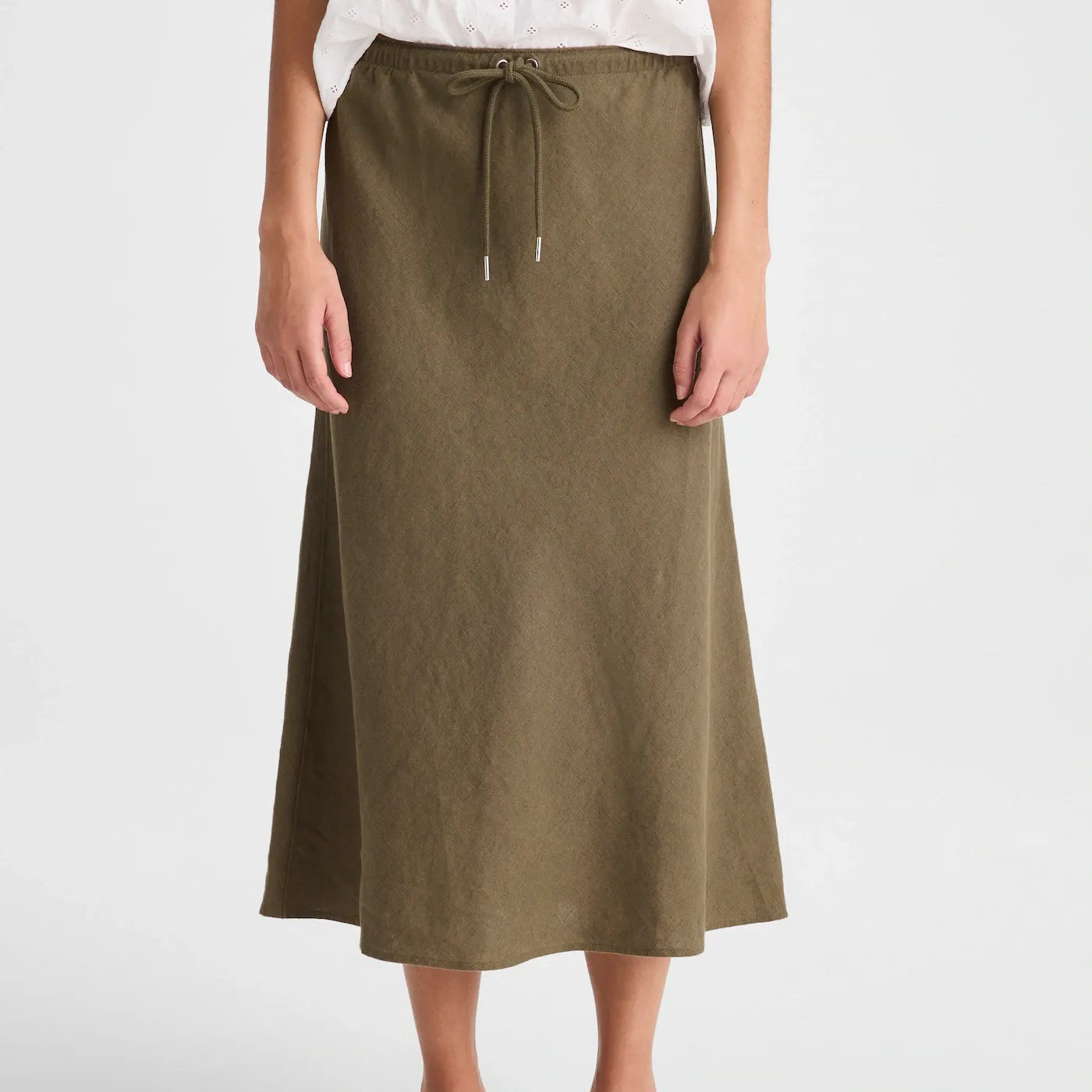 A woman wearing an olive green midi skirt with a drawstring waist, paired with a white top and black flip-flop sandals, shown against a light gray studio background.
