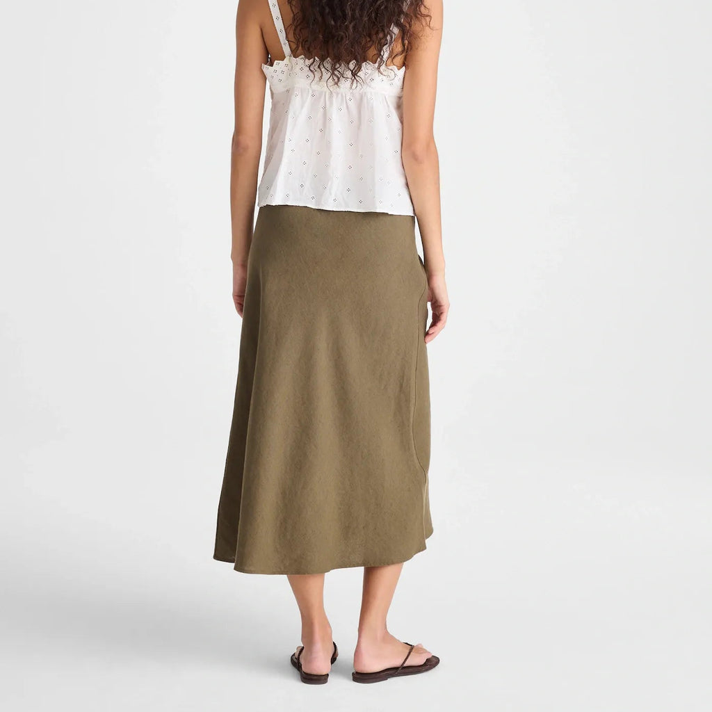 A woman shown from the back wearing a white sleeveless top with thin straps and a long olive green skirt, standing against a light gray studio background.