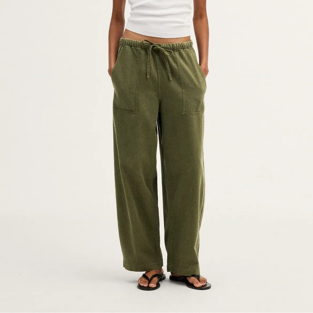 A model stands against a plain light background wearing olive green wide-leg drawstring pants, a white short-sleeve cropped T-shirt, and dark sandals.