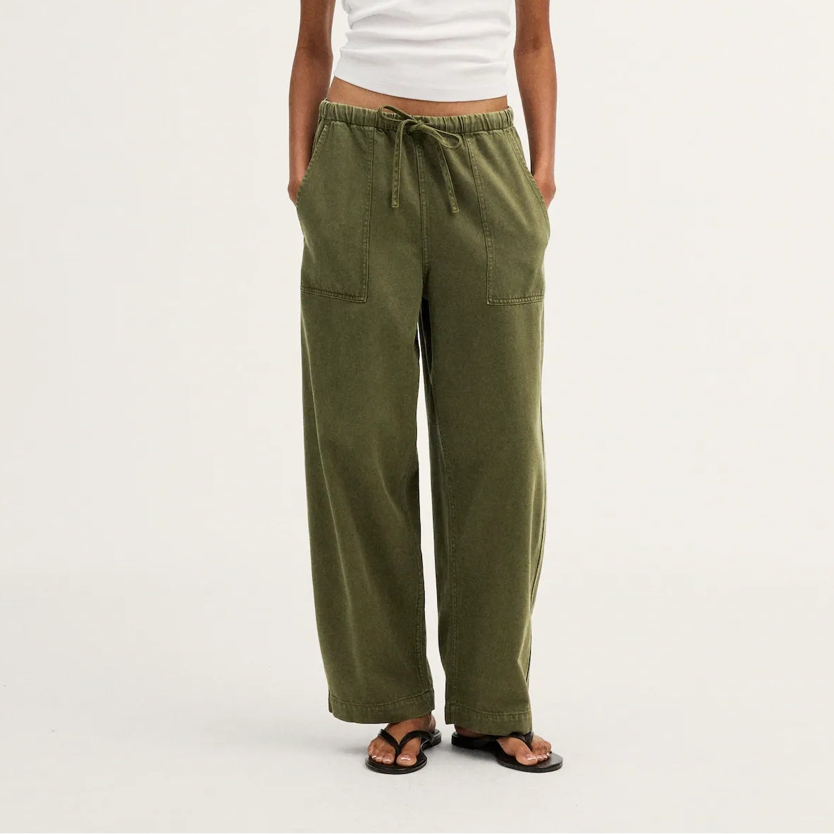 A model stands against a plain light background wearing olive green wide-leg drawstring pants, a white short-sleeve cropped T-shirt, and dark sandals.