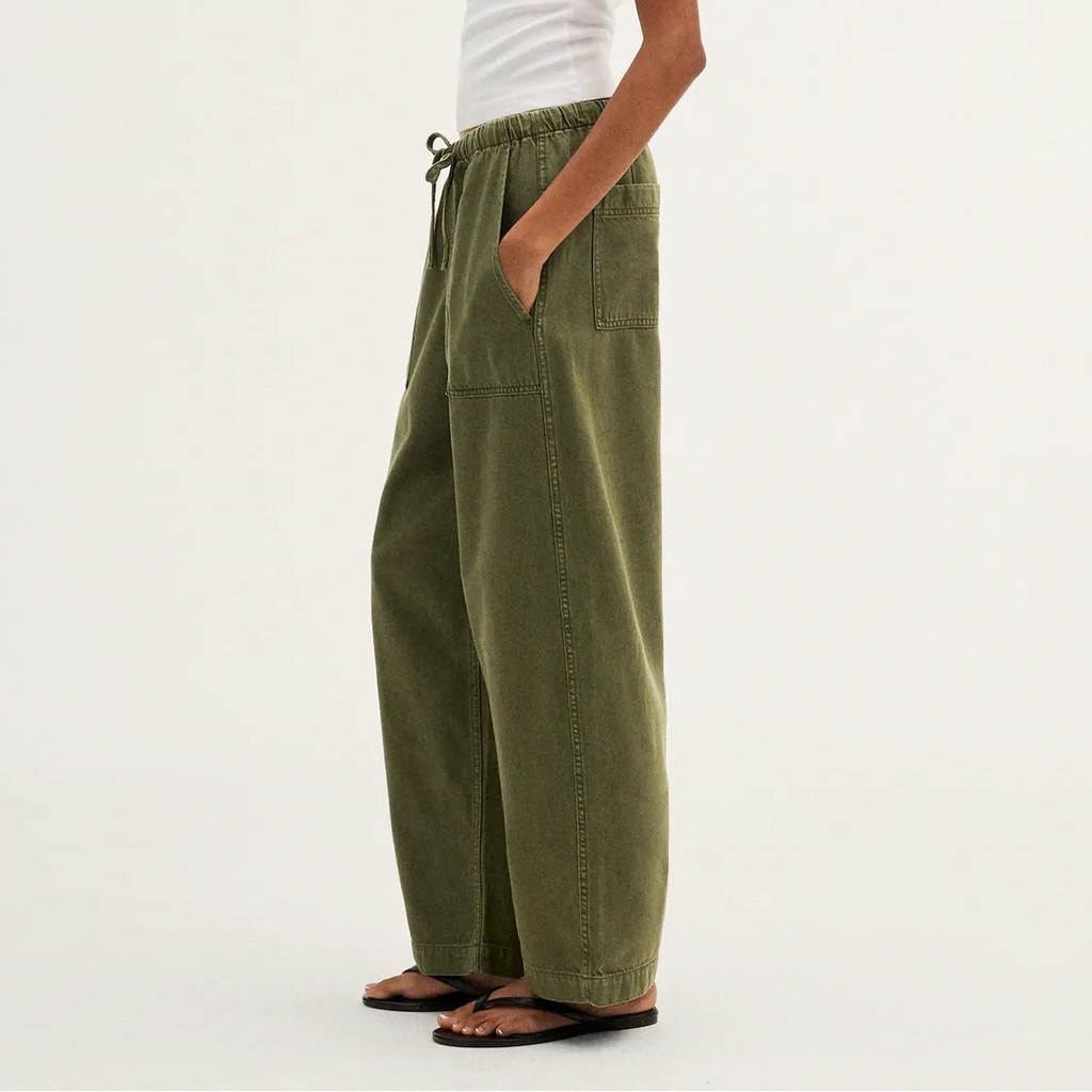 A model wearing olive green wide-leg corduroy pants, a white t-shirt, and brown sandals, shown in profile against a plain white studio background.