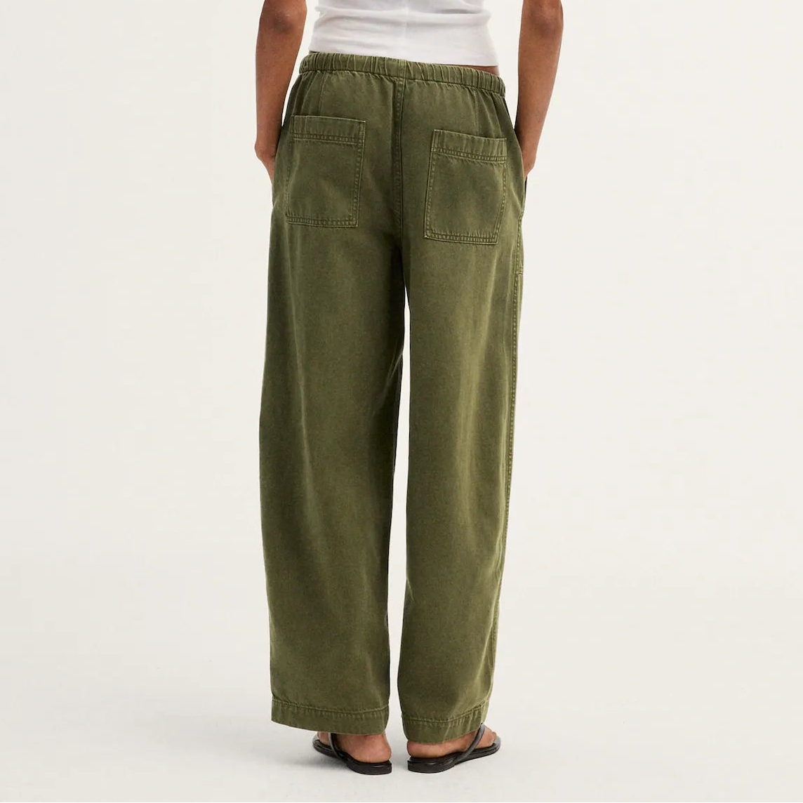 A model shown from the back wearing olive green wide-leg pants, a white short-sleeve t-shirt, and black sandals against a plain off-white studio background.