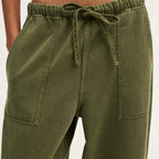 Close-up of a person wearing olive green drawstring pants with side pockets, paired with a white cropped top against a light neutral background.