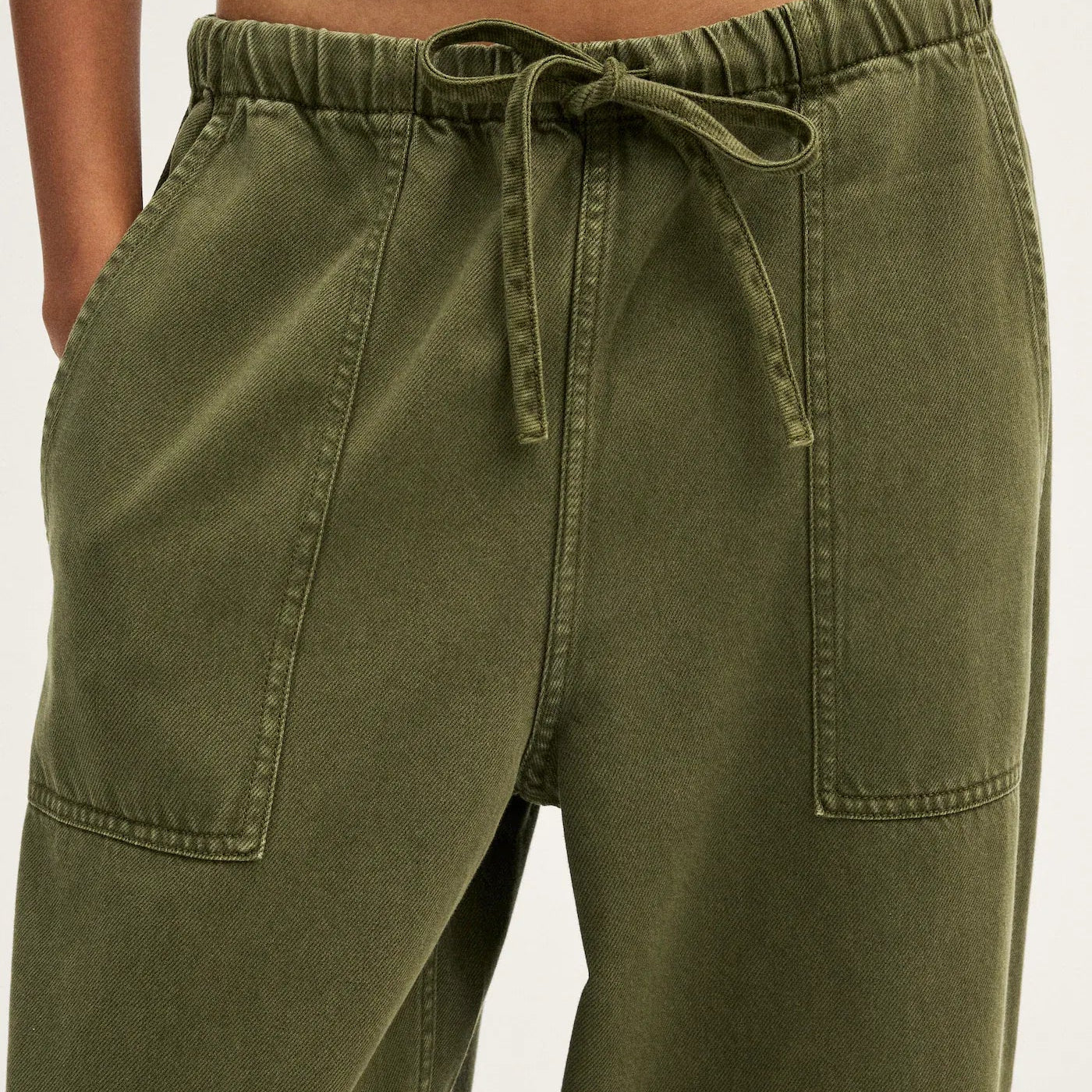Close-up of a person wearing olive green drawstring pants with side pockets, paired with a white cropped top against a light neutral background.