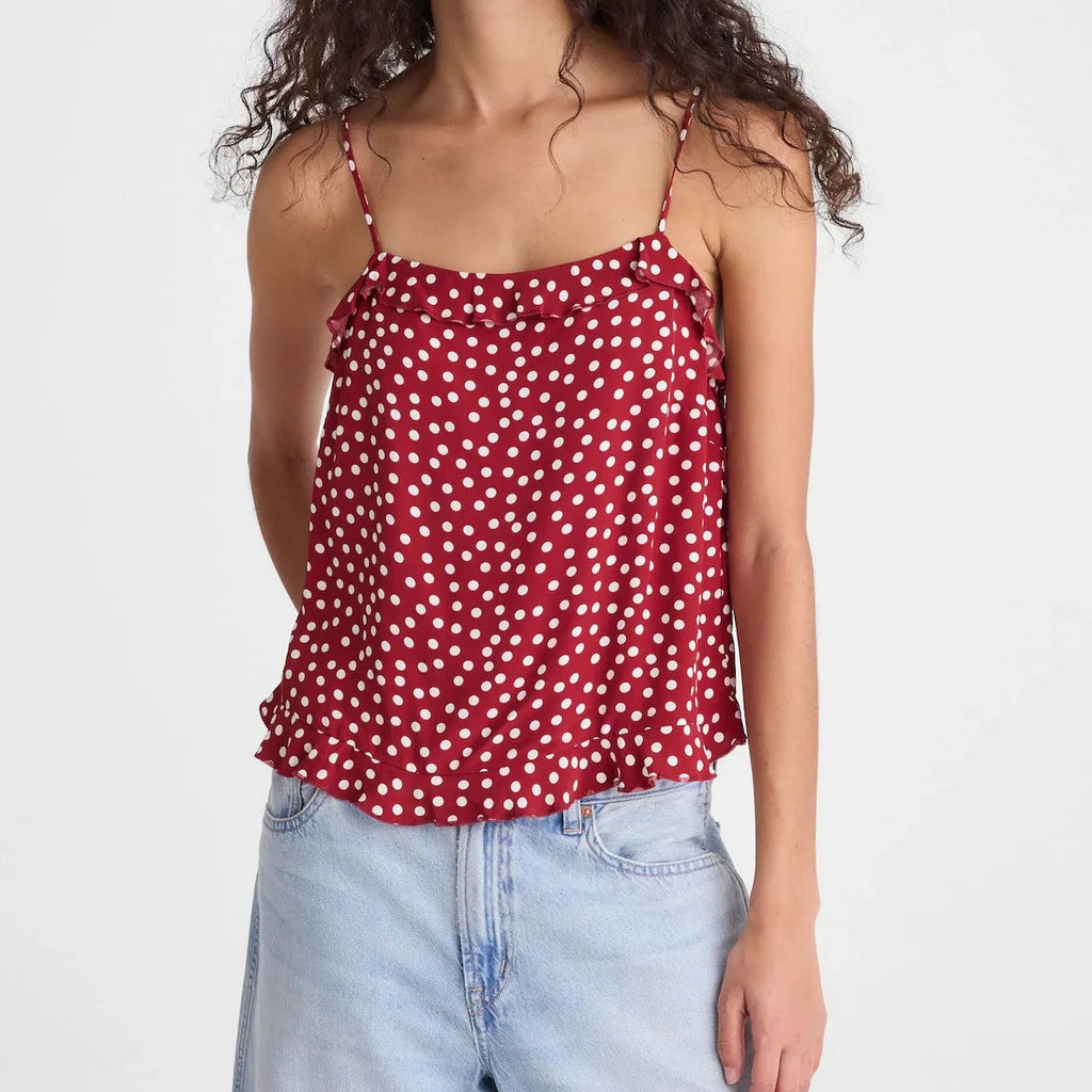 A woman with long, curly dark hair wearing a red spaghetti-strap camisole top with a white polka-dot pattern and light-wash blue jeans, standing against a solid light gray background.
