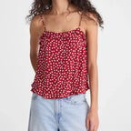 A woman with long, curly dark hair wearing a red spaghetti-strap camisole top with a white polka-dot pattern and light-wash blue jeans, standing against a solid light gray background.