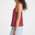 A woman with long, curly dark hair is shown from a side-back angle wearing a red camisole top with white polka dots and light-wash denim jeans against a plain white studio background.