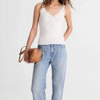 A woman modeling a white lace-trimmed camisole top, light-wash wide-leg jeans, and brown suede loafers, holding a small tan leather crossbody bag against a plain light gray studio background.