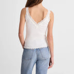 A woman viewed from the back wearing a white lace-trimmed camisole top and light blue jeans against a plain light gray studio background.