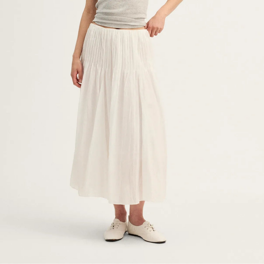 A woman modeling a light gray ribbed tank top and a long, pleated off-white midi skirt, paired with white sneakers, against a plain white studio background.