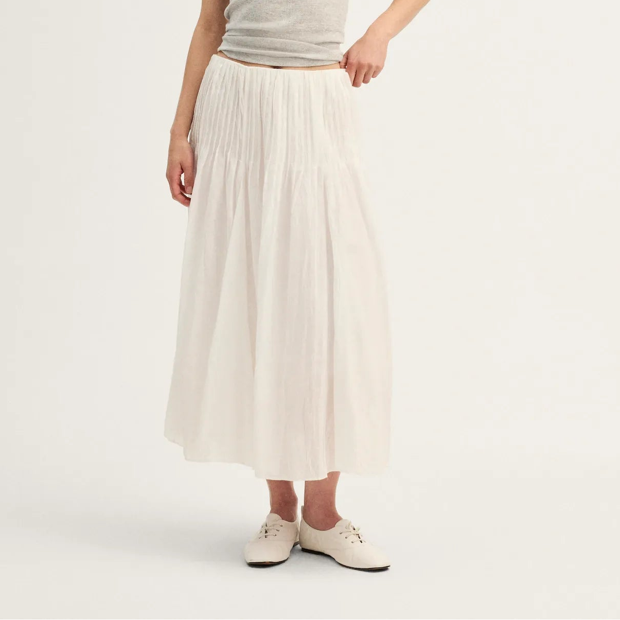 A woman modeling a light gray ribbed tank top and a long, pleated off-white midi skirt, paired with white sneakers, against a plain white studio background.