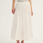A model wearing a high-waisted, pleated white midi skirt with a textured knit top and white lace-up shoes, shown against a plain white studio background.