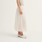 A woman stands in profile wearing a light gray sleeveless top and a long, flowing off-white skirt, paired with white shoes, against a plain light background.