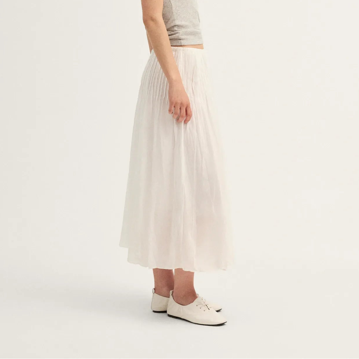 A woman stands in profile wearing a light gray sleeveless top and a long, flowing off-white skirt, paired with white shoes, against a plain light background.
