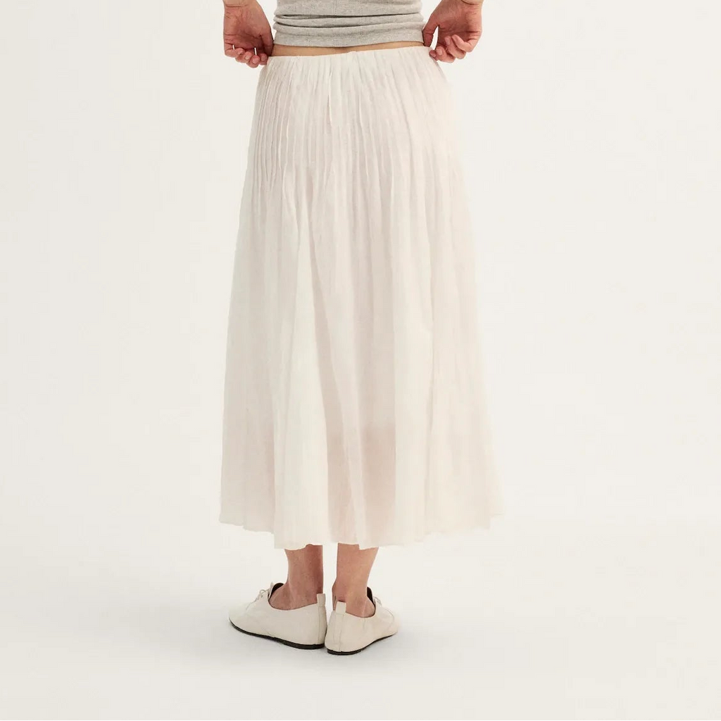A woman shown from the back wearing a light gray ribbed tank top and a long, flowing off-white pleated midi skirt, paired with white slip-on shoes, against a plain white studio background.