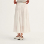 A woman shown from the back wearing a light gray ribbed tank top and a long, flowing off-white pleated midi skirt, paired with white slip-on shoes, against a plain white studio background.