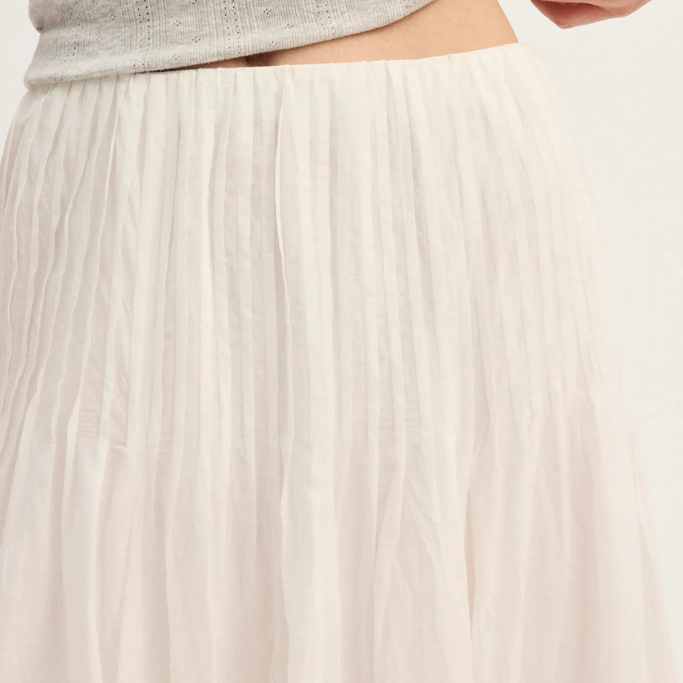 Close-up of a person wearing a light heather gray ribbed knit top and an off-white pleated skirt, lifting the hem of the top to show the waistband.